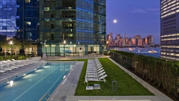 A modern hotel pool area with a city skyline view, a grassy lawn, and lounge chairs. - Green Suites (Jersey City)