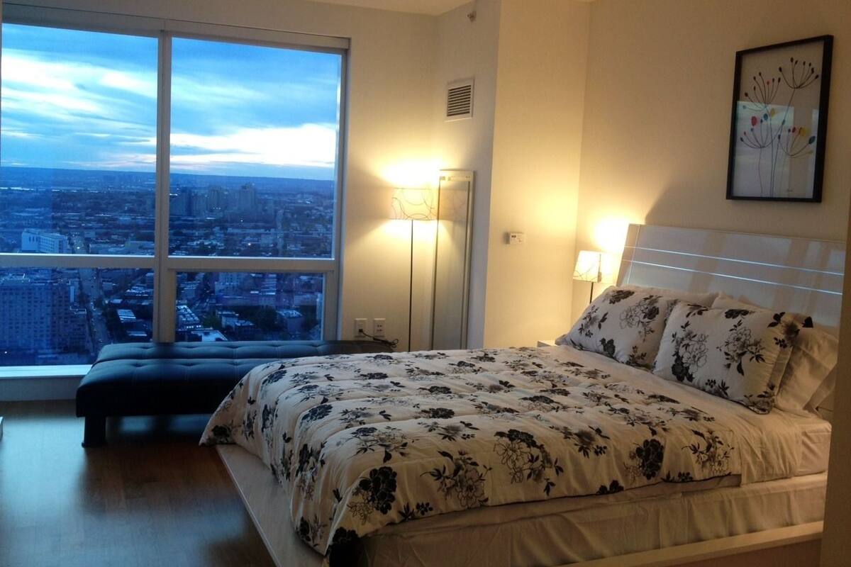 A bedroom with a large window, a bed with floral bedding, a bedside table, and a nightstand.