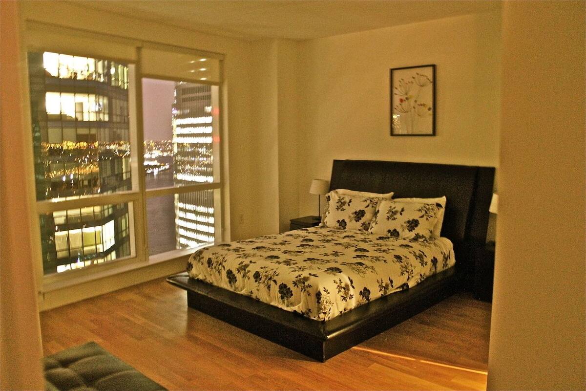 A bedroom with a large window, a bed with a floral bedspread, a nightstand, and a framed picture on the wall.
