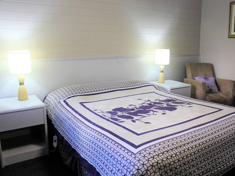 Una camera d'albergo con un letto rifatto, due lampade sui comodini, una sedia e un quadro appeso al muro.