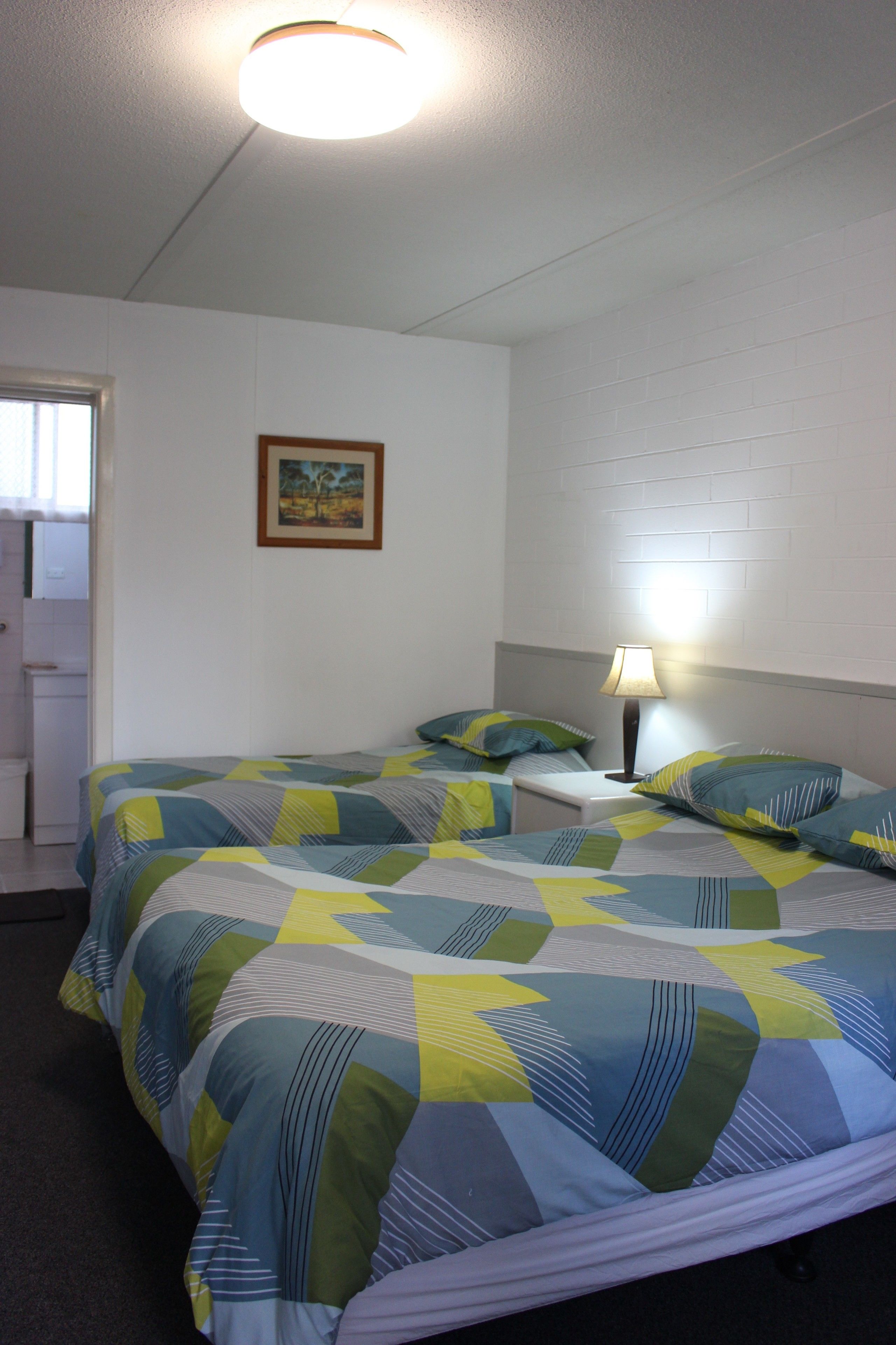 Habitación de hotel con dos camas, una mesita de noche con lámpara, un cuadro en la pared y una puerta a la izquierda.