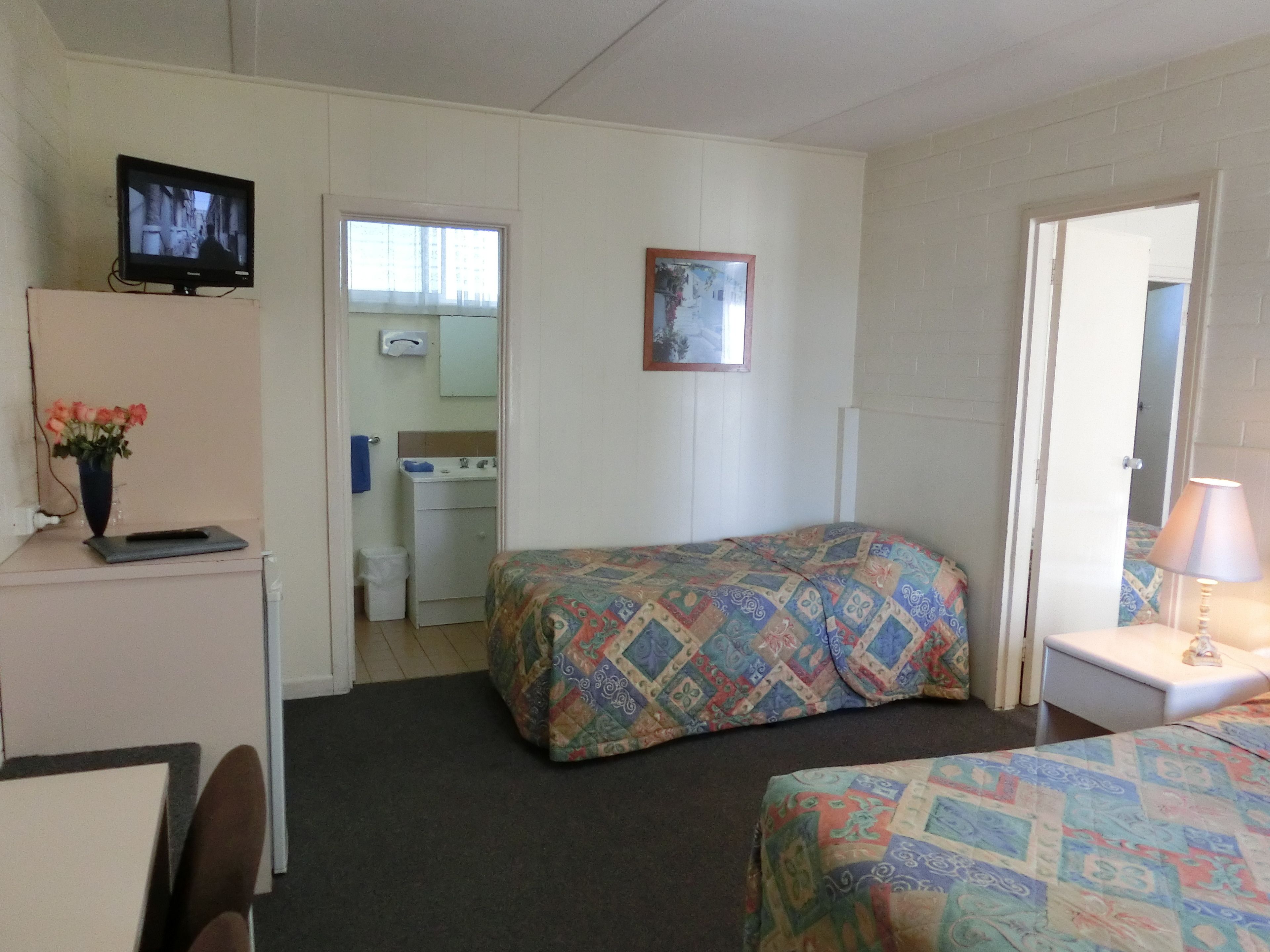 A small, simple room with two beds, a desk, a TV, and a bathroom visible through an open door.