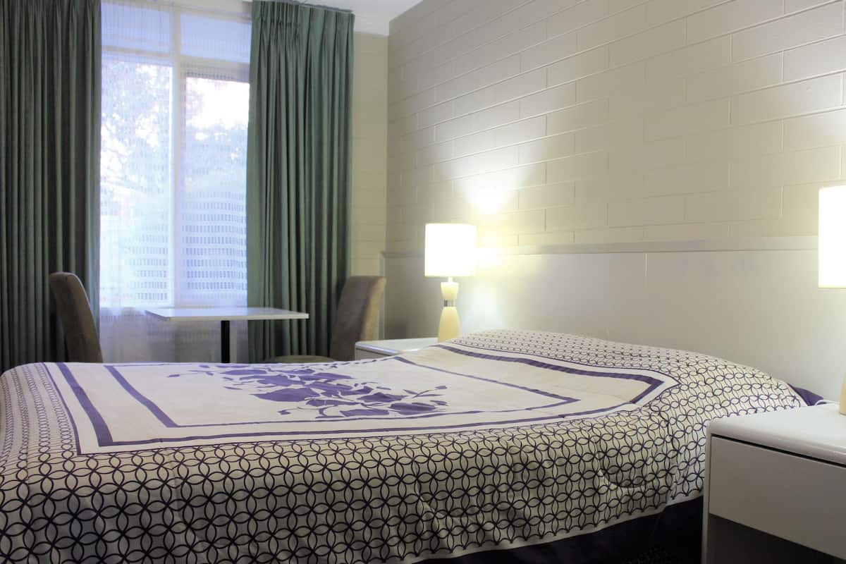 A hotel room with a bed, two bedside lamps, a chair, a table, and a window with curtains.