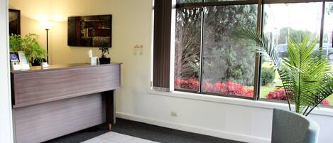 A reception area with a counter, a chair, a lamp, and a window with a view of trees and flowers.