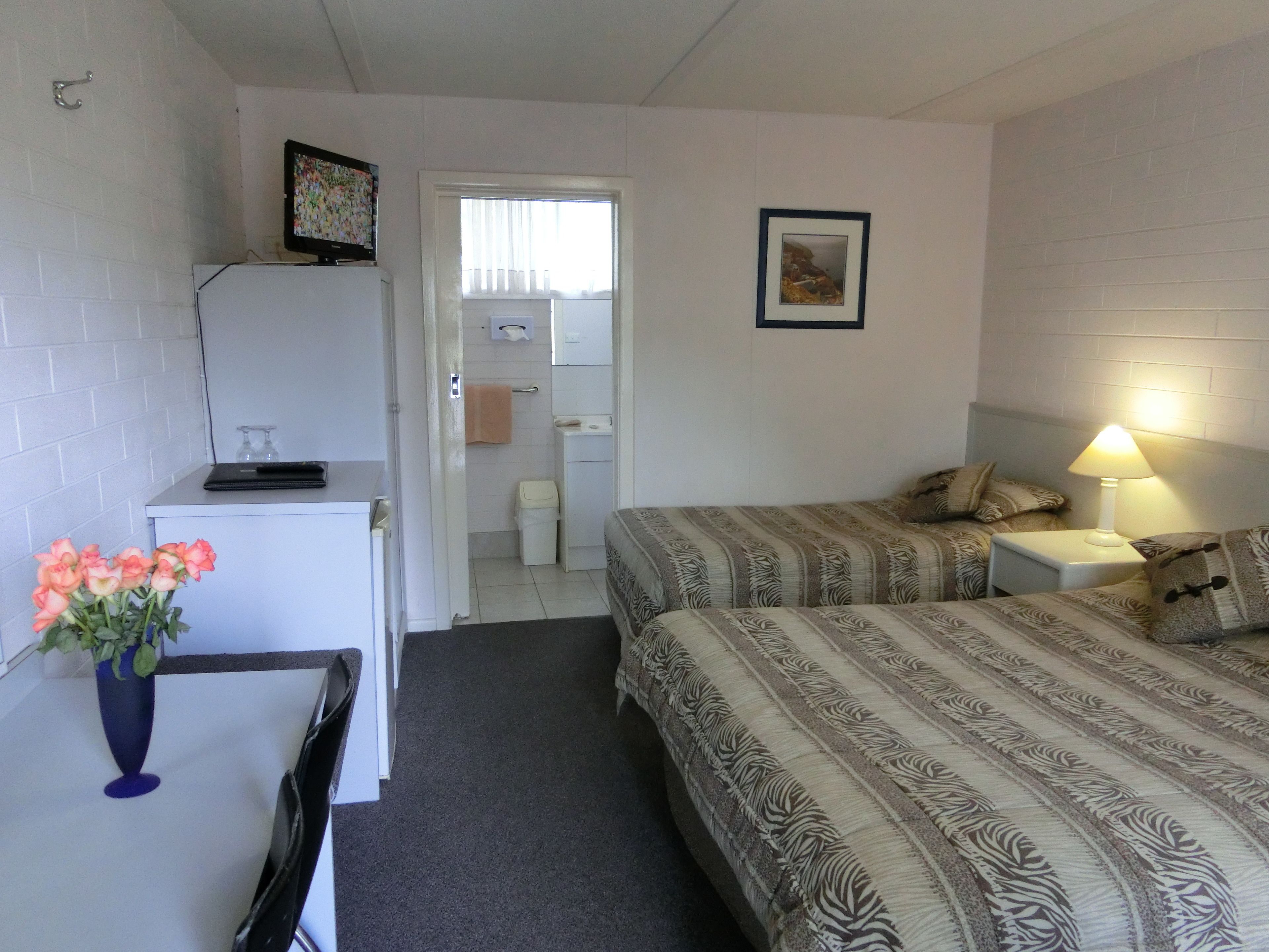 A hotel room with a bed, bedside table, lamp, and a desk with a computer and a vase of flowers.