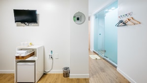A room with a white wall, a mounted TV, a small refrigerator, a trash bin, and a blue glass door leading to a bathroom.