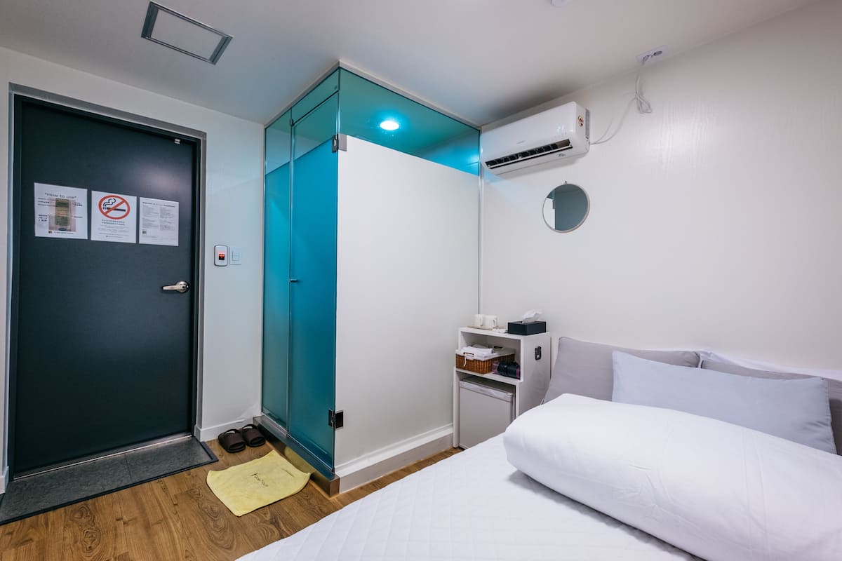 Habitació d'hotel amb un llit, una petita taula de nit, aire condicionat fixat a la paret, una porta amb un panell de vidre i un parell de xalets al terra.