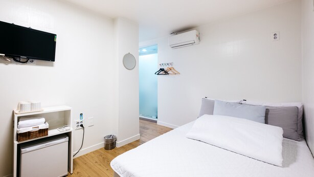 A hotel room with a bed, a small desk with a flat-screen TV, a mini-fridge, a basket, and a hanging clothes rack.