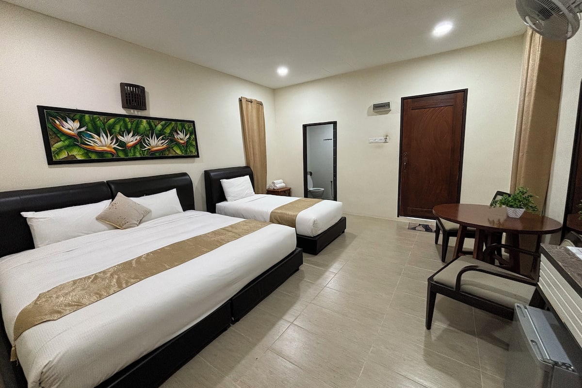 A hotel room with two beds, a small table, a chair, a potted plant, a painting, and a door.