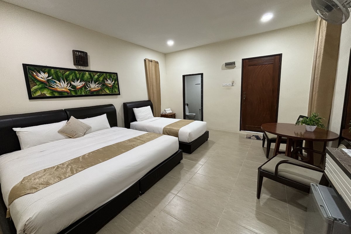 A hotel room with two beds, a dining table, a chair, a potted plant, a painting, and a mirror.