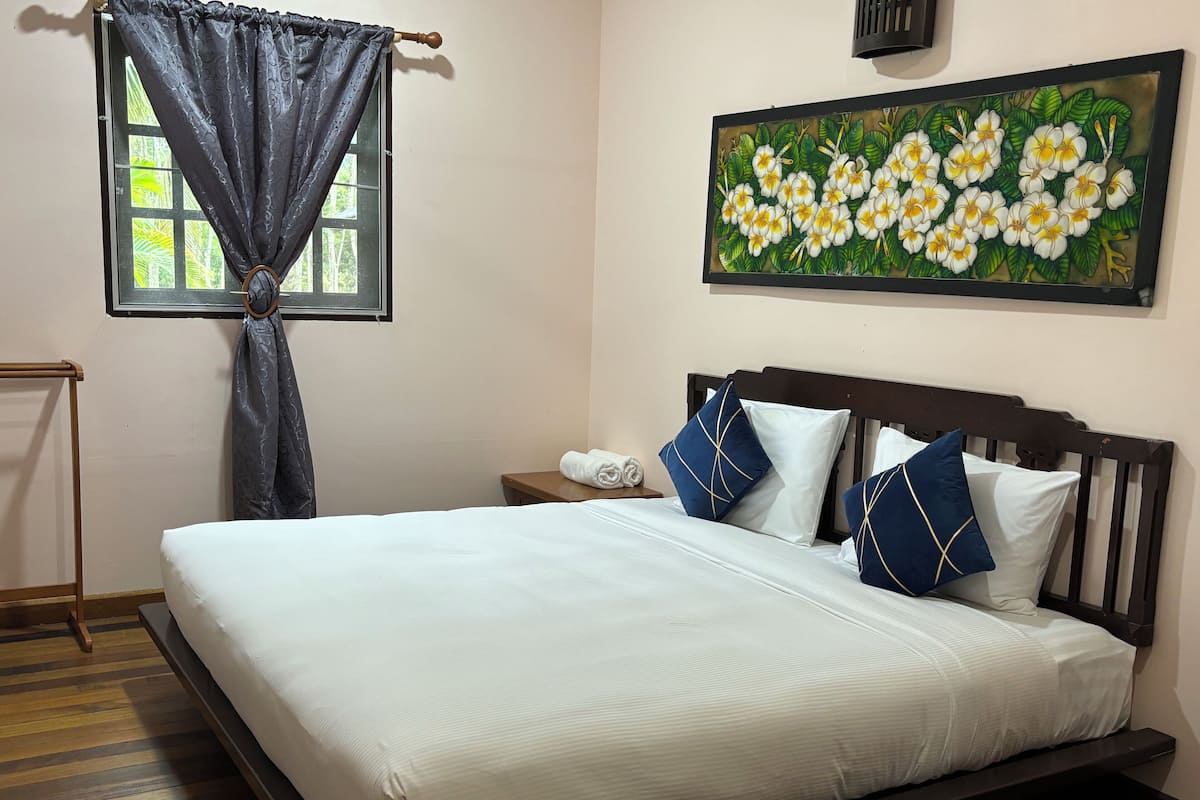 A neatly made bed with white bedding and blue and white pillows, a wooden headboard, a small bedside table with a lamp, and a window with curtains.