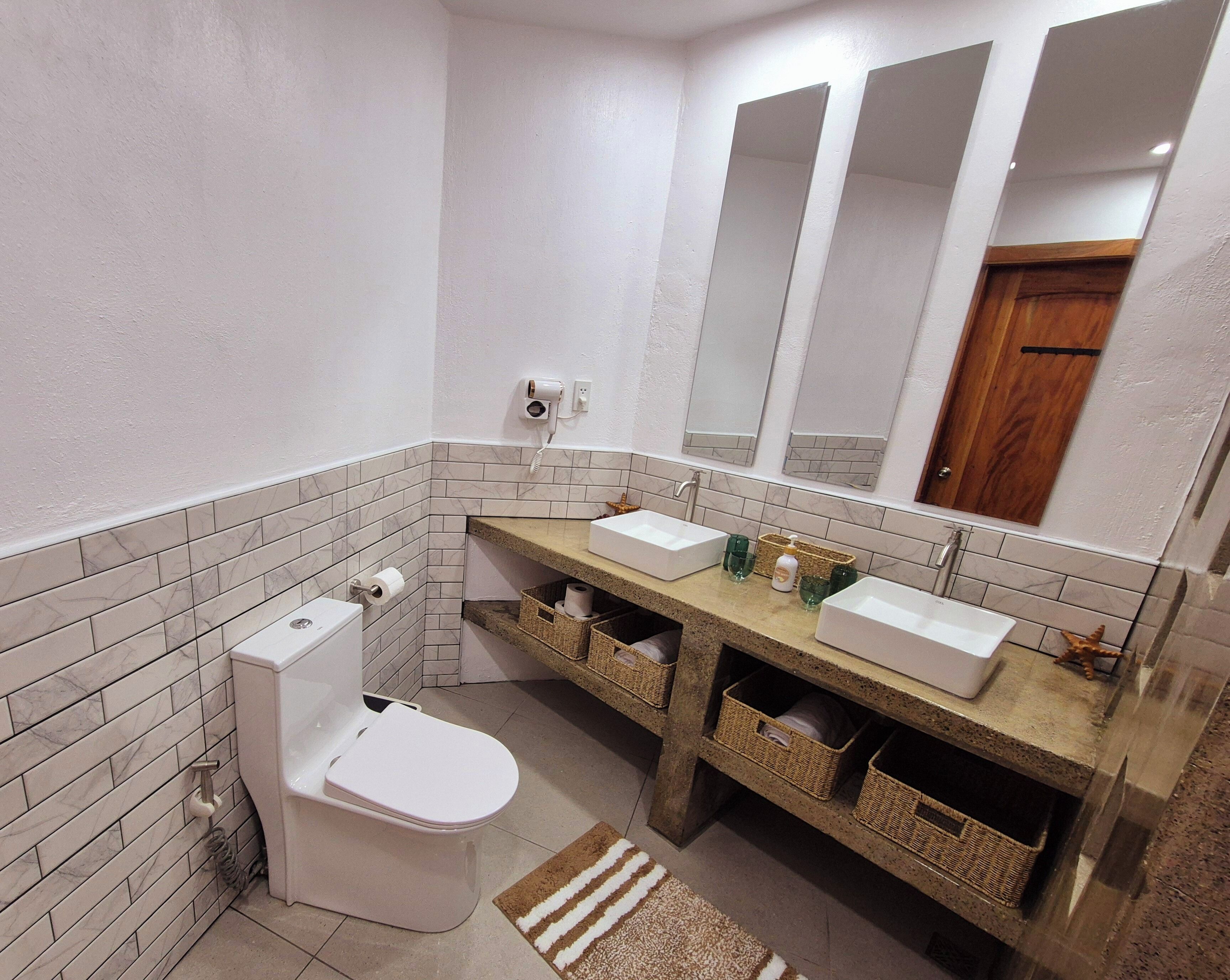 A bathroom with two sinks, a toilet, and a mirror. There are wicker baskets under the sinks and a roll of toilet paper on the wall.