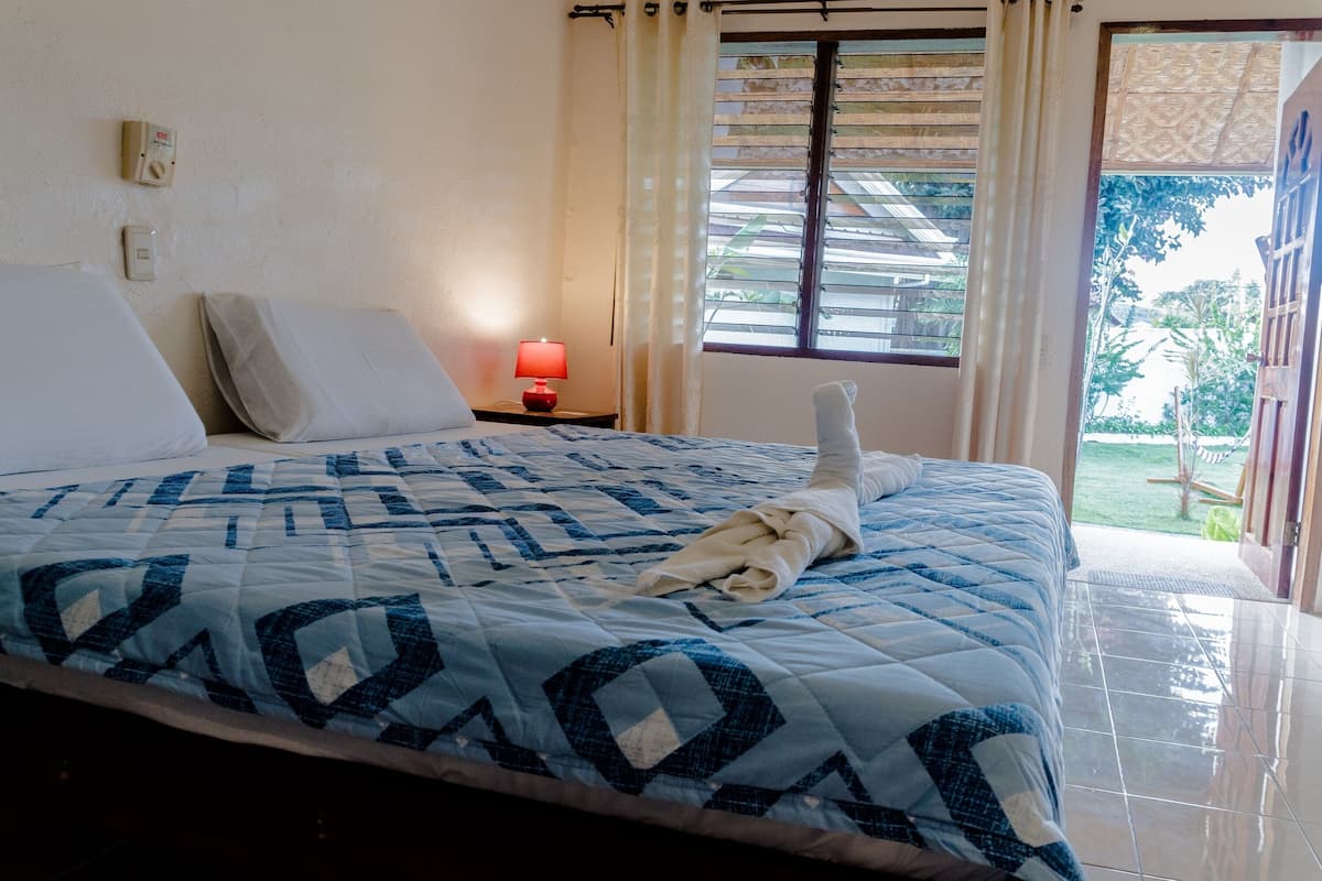 Een bed met een blauwe sprei, een rode lamp op een nachtkastje en een raam met jaloezieën.