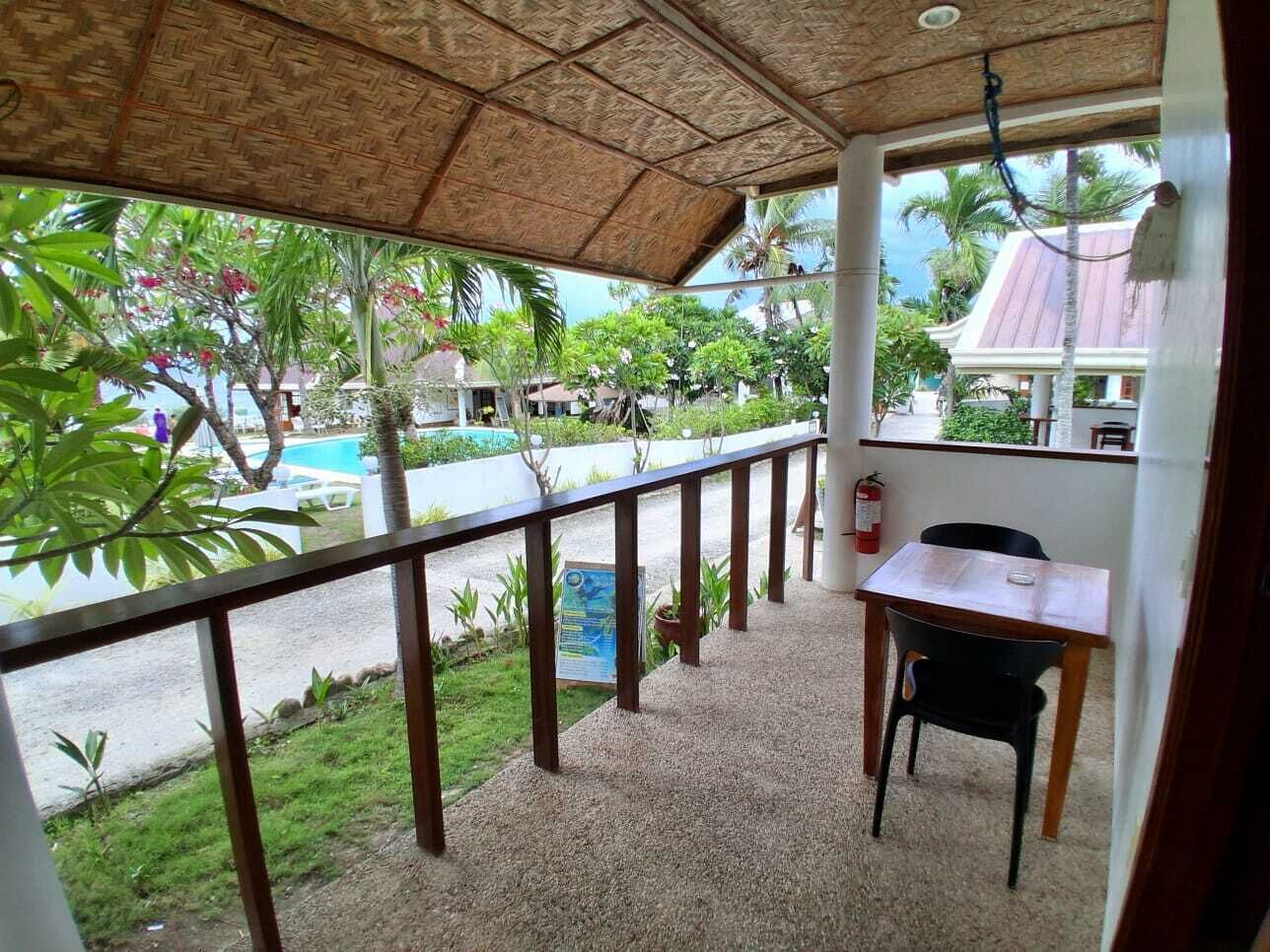 A covered balcony with a table and chair, a fire extinguisher, and a view of a swimming pool and greenery.