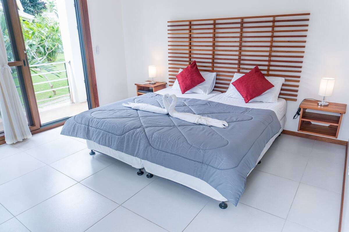 Een netjes opgemaakt bed met een grijze dekbedovertrek, twee rode kussens en een witte handdoek in de vorm van een zwaan. Er staan twee nachtkastjes met lampen, en een houten latten hoofdgreep.