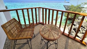 A balcony with two wicker chairs and a small round table, overlooking a clear blue sea and distant mountains.
