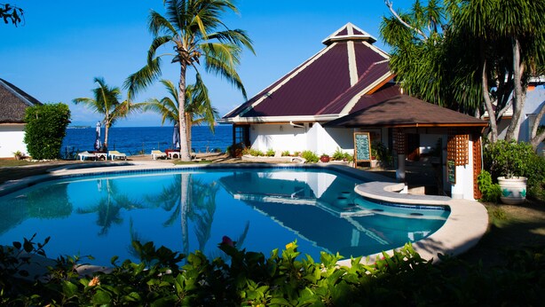 A pool area with a view of the ocean, surrounded by palm trees and a building with a red roof.