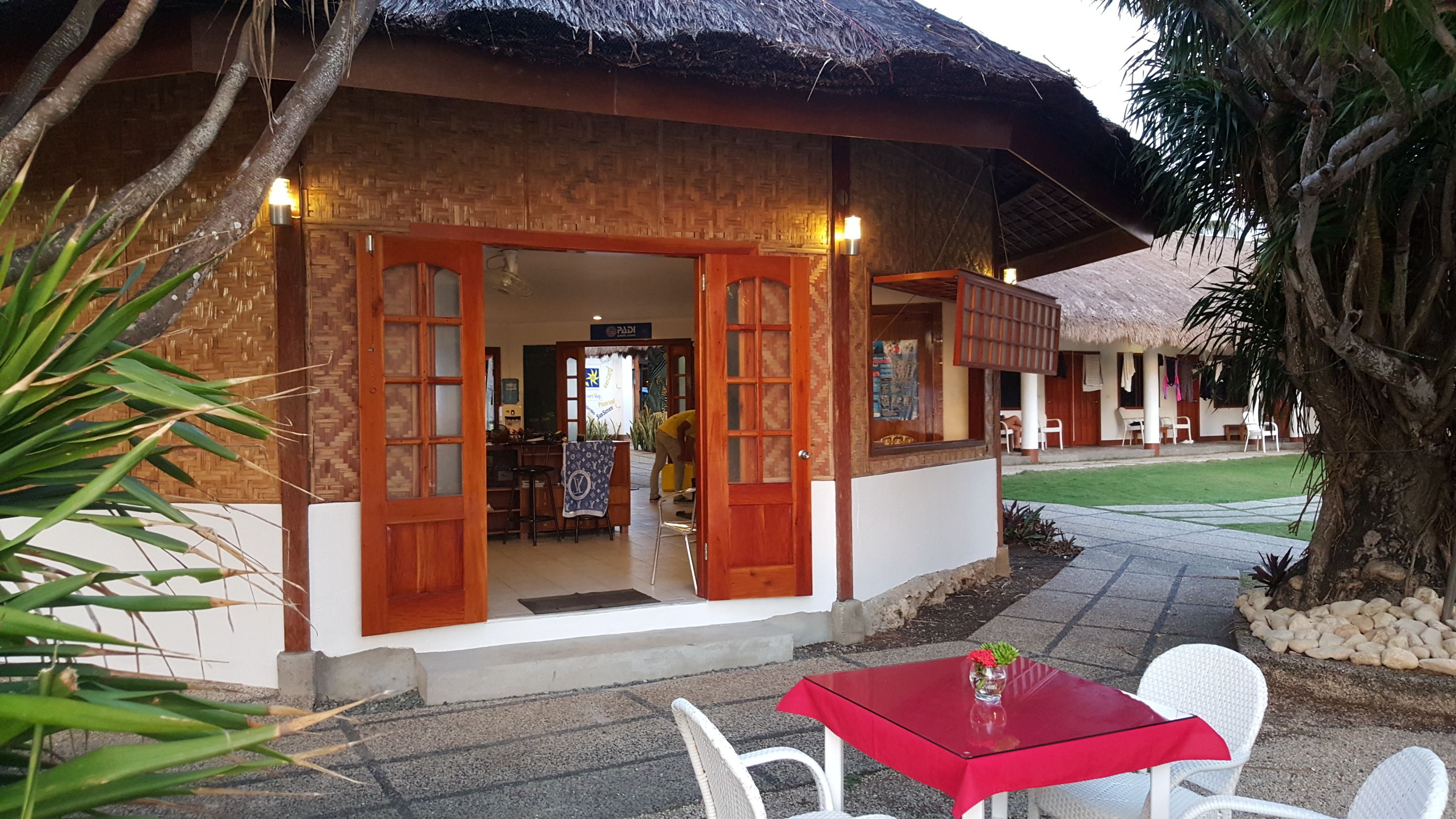 A thatched-roof building with wooden doors and windows, a small outdoor seating area with a red table and white chairs, and a well-maintained lawn.