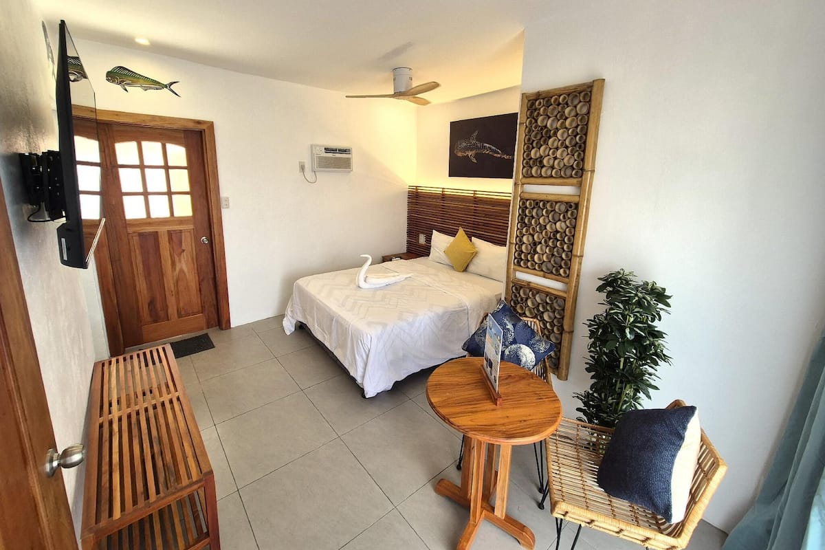 Een hotelkamer met een bed, een klein houten tafeltje, een stoel, een kamerplant en een decoratief paneel aan de muur.