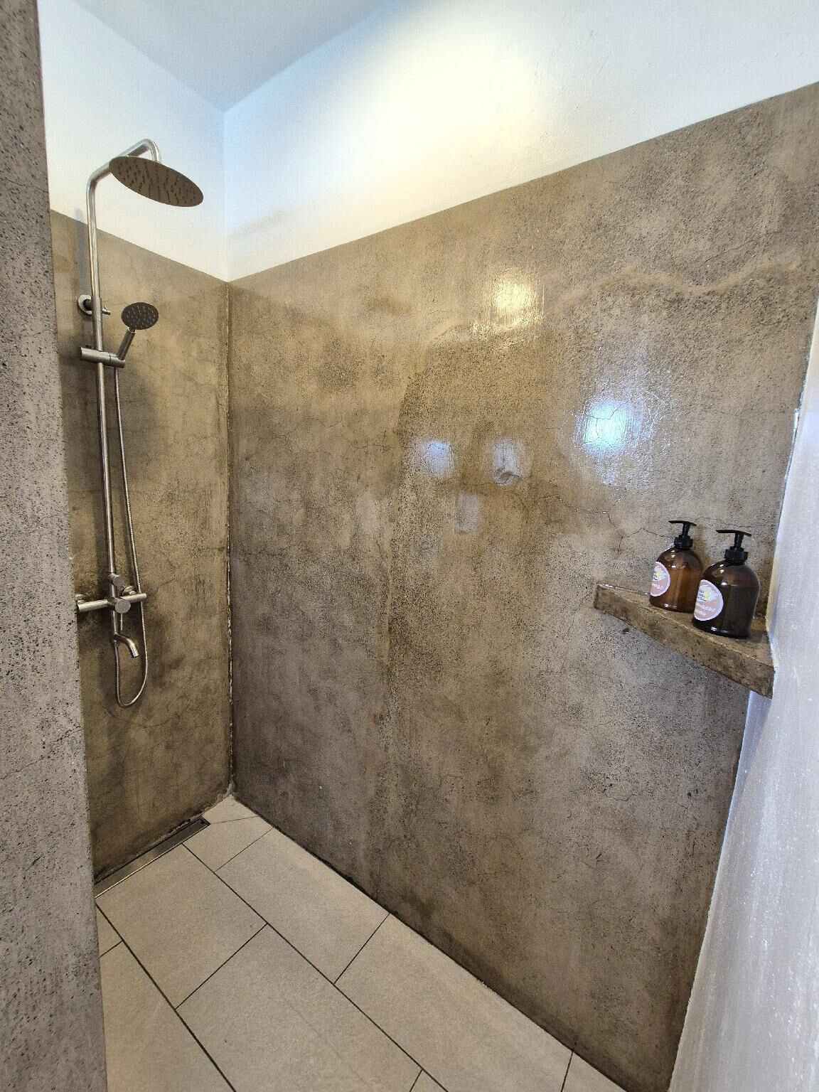 Douchegedeelte met een handdouche en een douchekop aan de muur, een betegelde vloer en een plank met flessen.