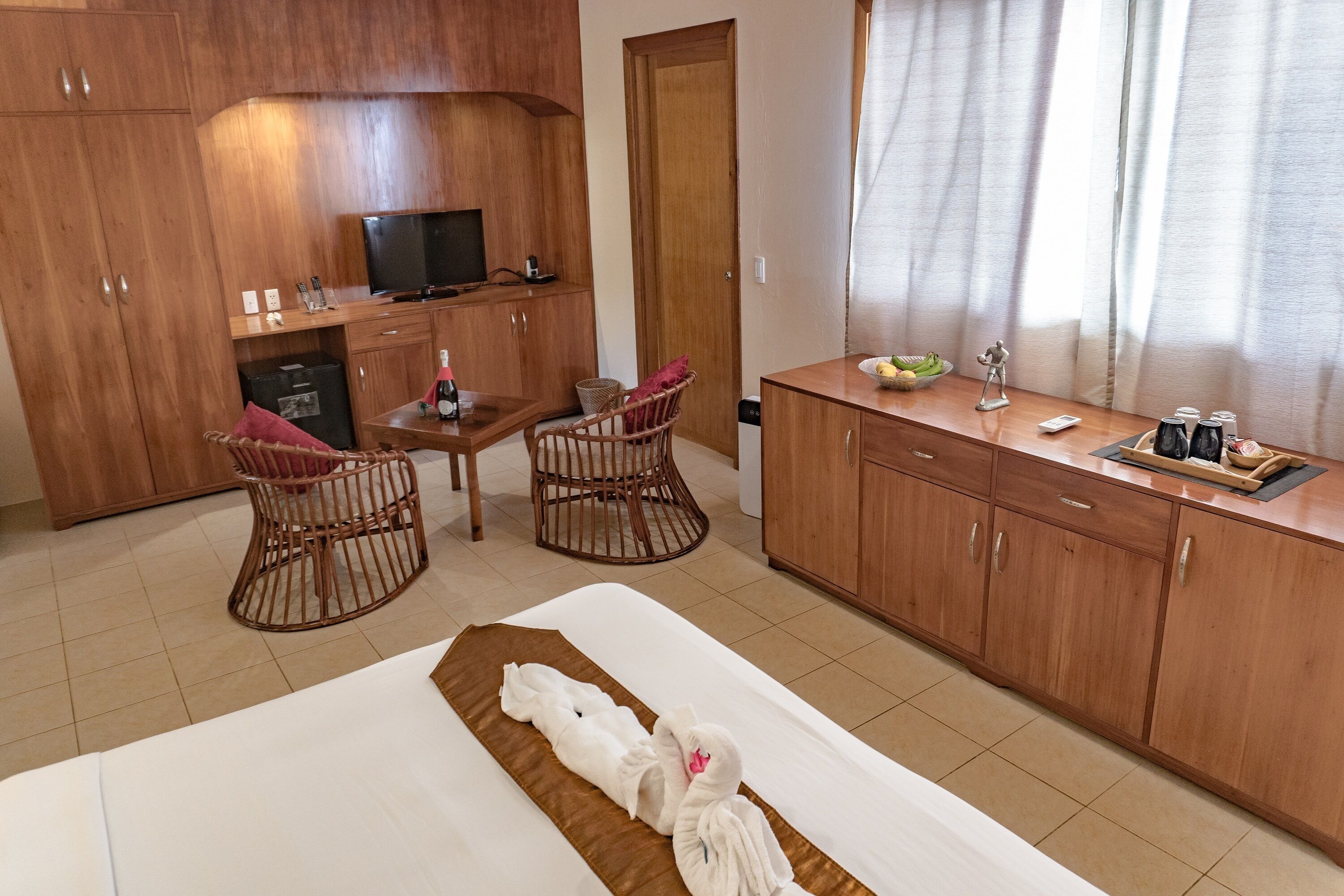 Une chambre d’hôtel avec un lit, un bureau en bois, deux fauteuils en osier, une télévision et une fenêtre avec des rideaux.