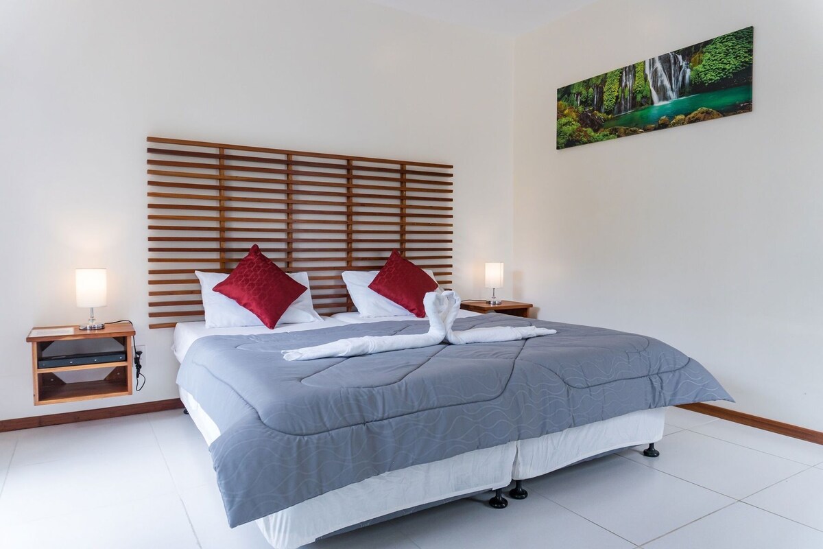 A neatly made bed with a grey duvet, two red pillows, and a white towel folded on the bed. There are two bedside tables with lamps, a wooden headboard, and a painting of a waterfall on the wall.