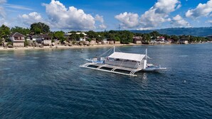 A traditional boat with a covered deck and a white canopy, floating on calm waters near a shoreline with houses and mountains in the background.