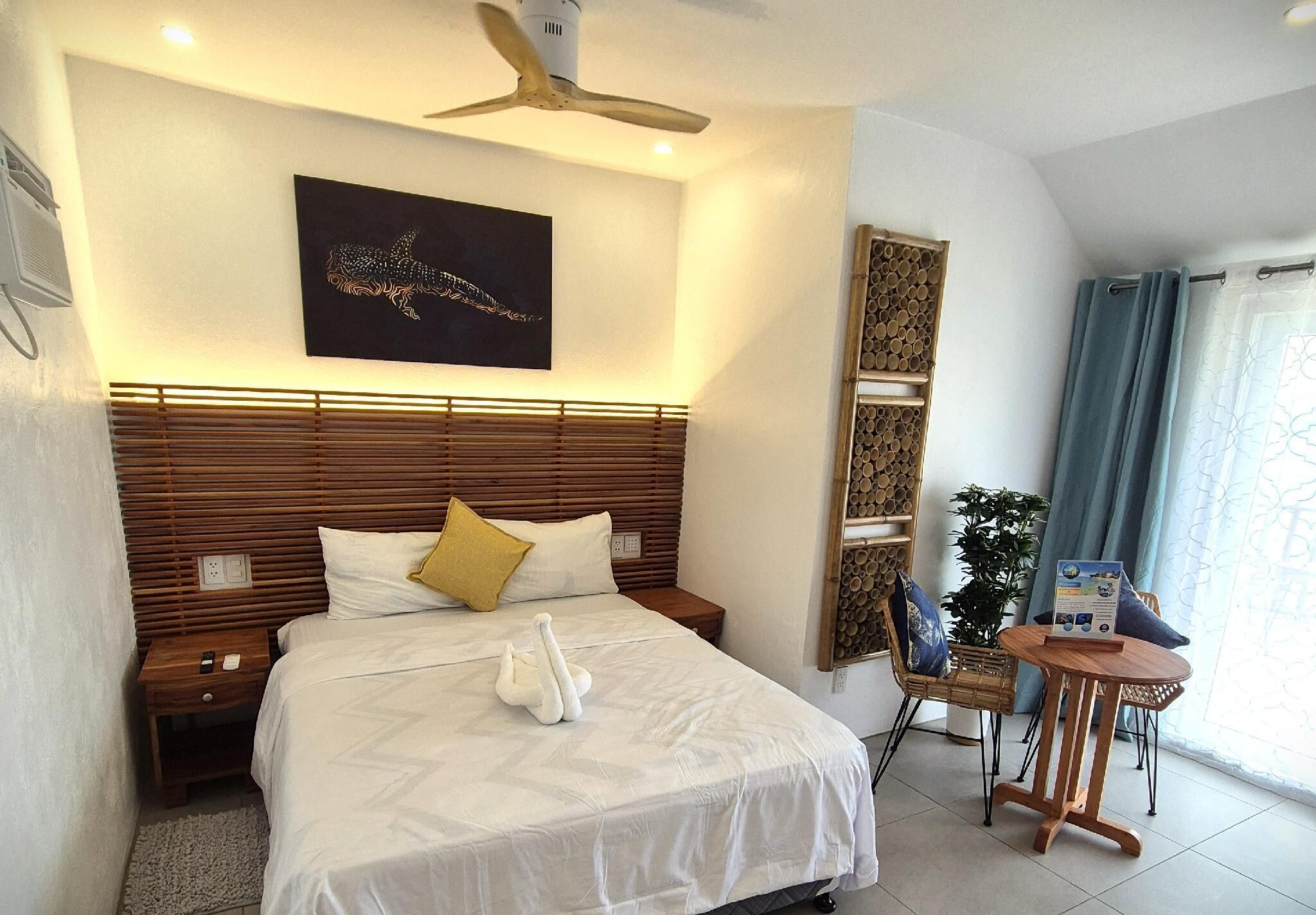 A hotel room with a large bed, wooden headboard, a ceiling fan, a wall-mounted artwork, a small table with a chair, and a potted plant.