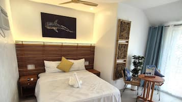 A hotel room with a large bed, wooden headboard, a ceiling fan, a wall-mounted artwork, a small table with a chair, and a potted plant.