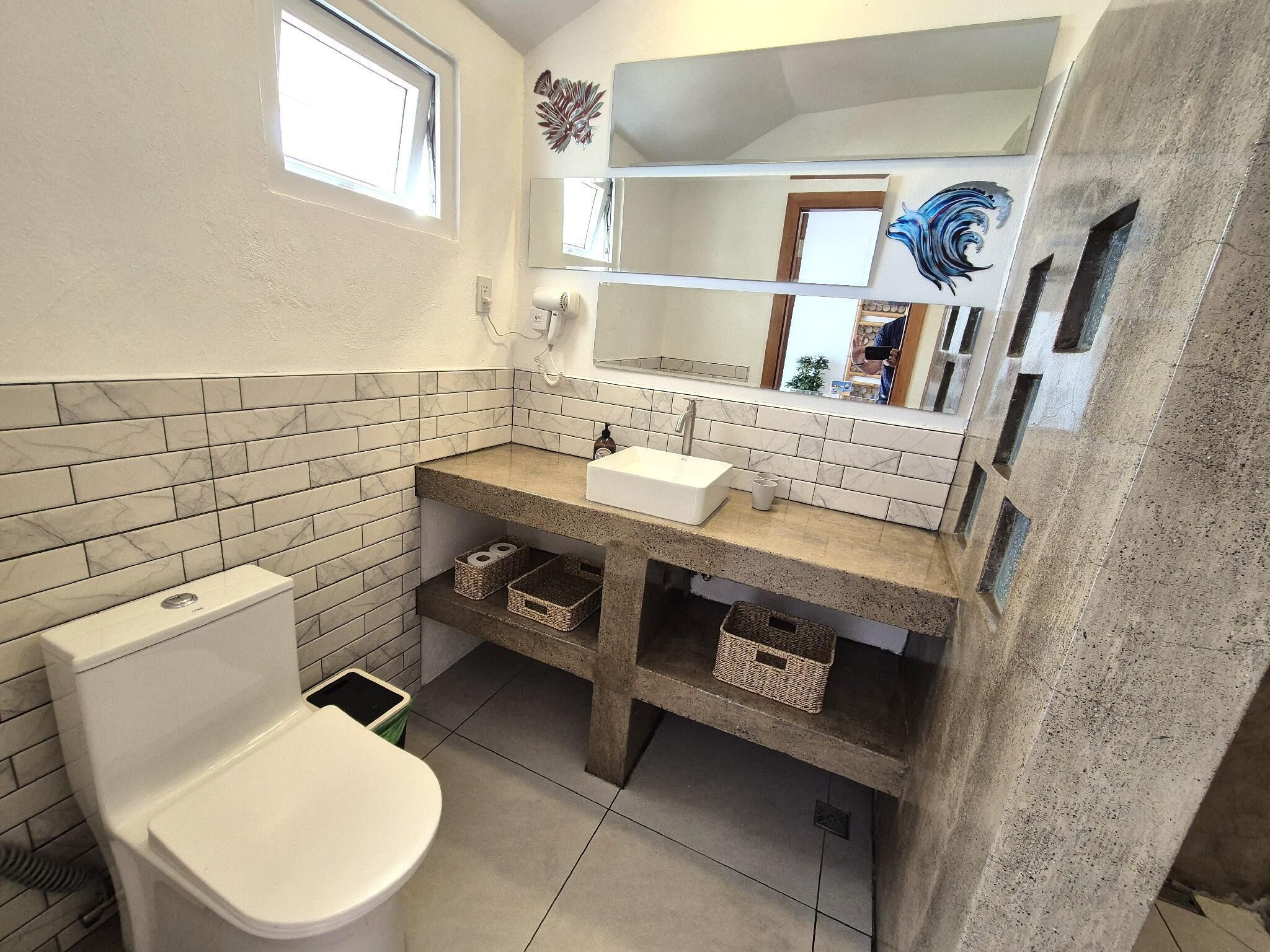 A bathroom with a toilet, a sink, and a mirror. There are baskets under the sink and a window above the toilet.