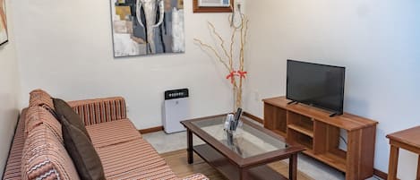 Una sala de estar con un sofá, una mesa de centro de vidrio, un televisor sobre un mueble de madera y un cuadro en la pared.