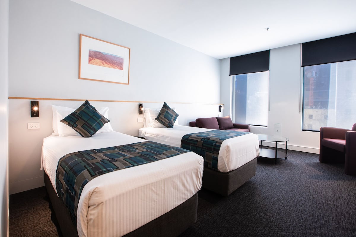 A hotel room with two beds, a small table, a chair, and a large window.