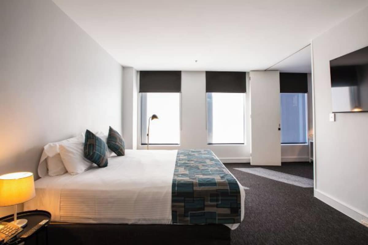 A hotel room with a bed, a nightstand, a lamp, and a view of the outside through large windows.
