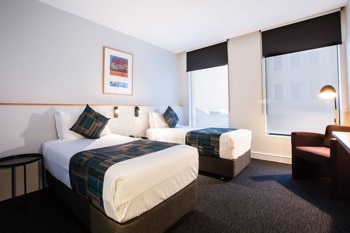 A hotel room with two beds, a desk, a chair, a framed picture on the wall, and a window with a view of buildings.