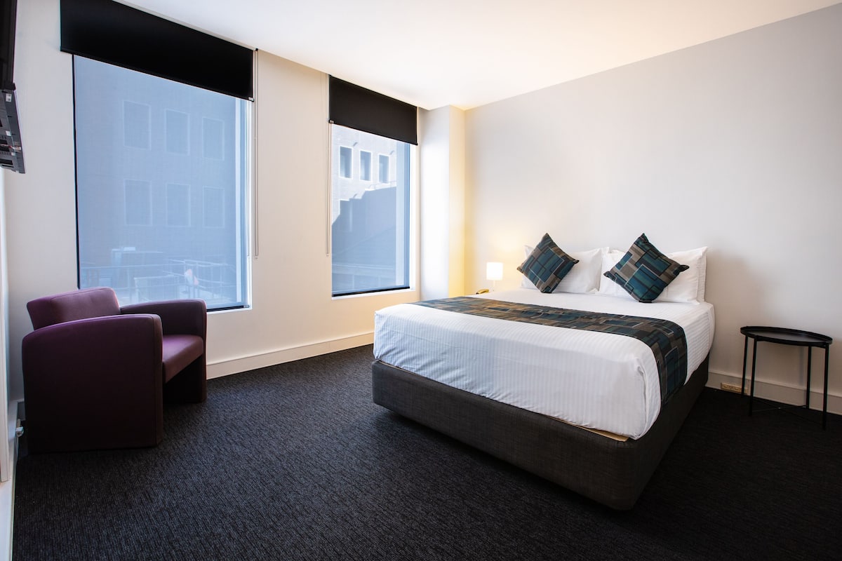 A hotel room with a large bed, two armchairs, a small table, and a window with a view of buildings.