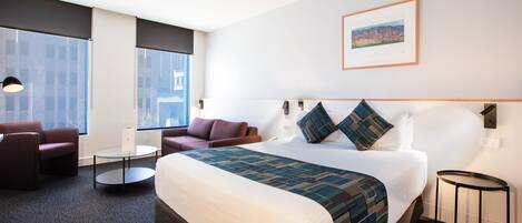 A hotel room with a large bed, two armchairs, a small table, and a large window.
