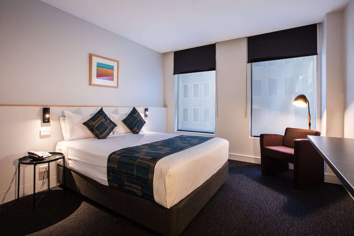 A hotel room with a large bed, a desk, and a chair. There is a window with a view of buildings outside.