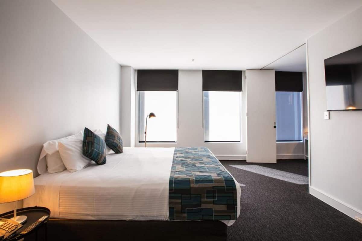 A hotel room with a bed, a desk with a lamp, and a view of the outside through large windows.