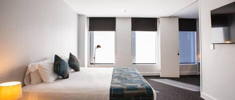 Quarto de hotel com cama, escrivaninha com abajur e vista exterior por grandes janelas.