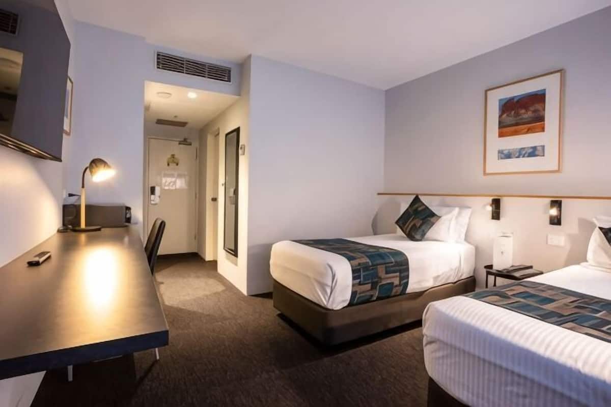 A hotel room with a desk, two beds, a chair, and a painting on the wall.