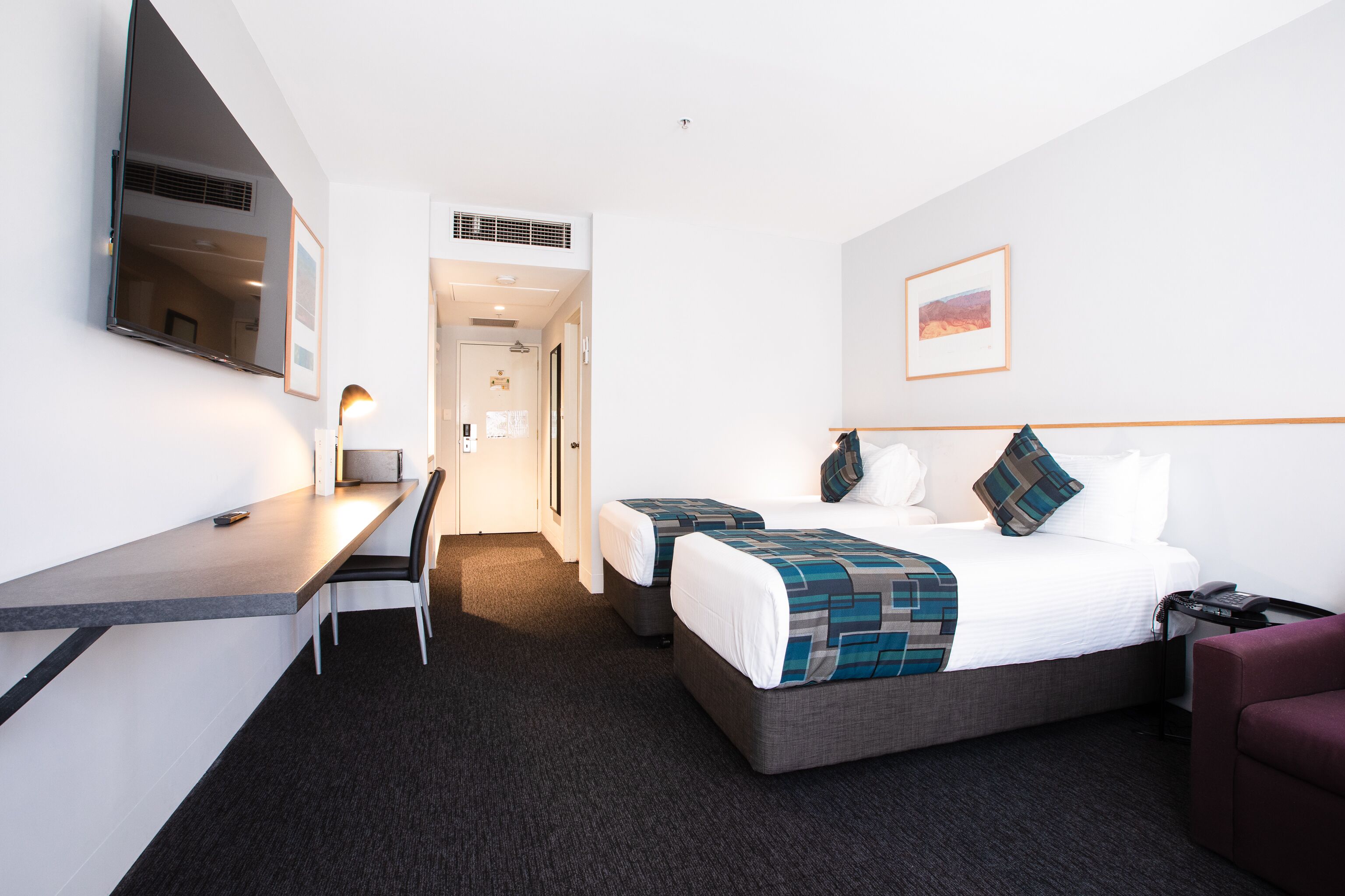 Een hotelkamer met een bed, bureau, stoel, televisie en een lijst aan de muur.