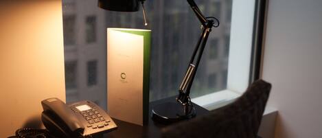 Un bureau avec un téléphone, une lampe et un objet vert, peut-être une plante, devant une fenêtre donnant sur des immeubles.