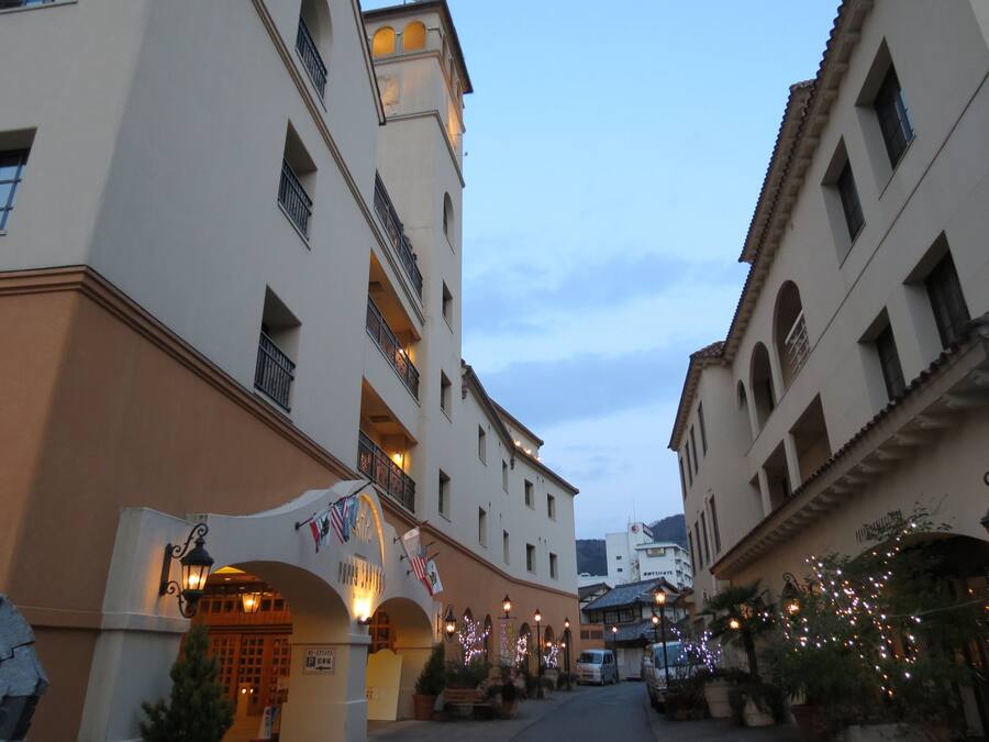 A narrow street flanked by buildings with balconies, street lamps, and flags.