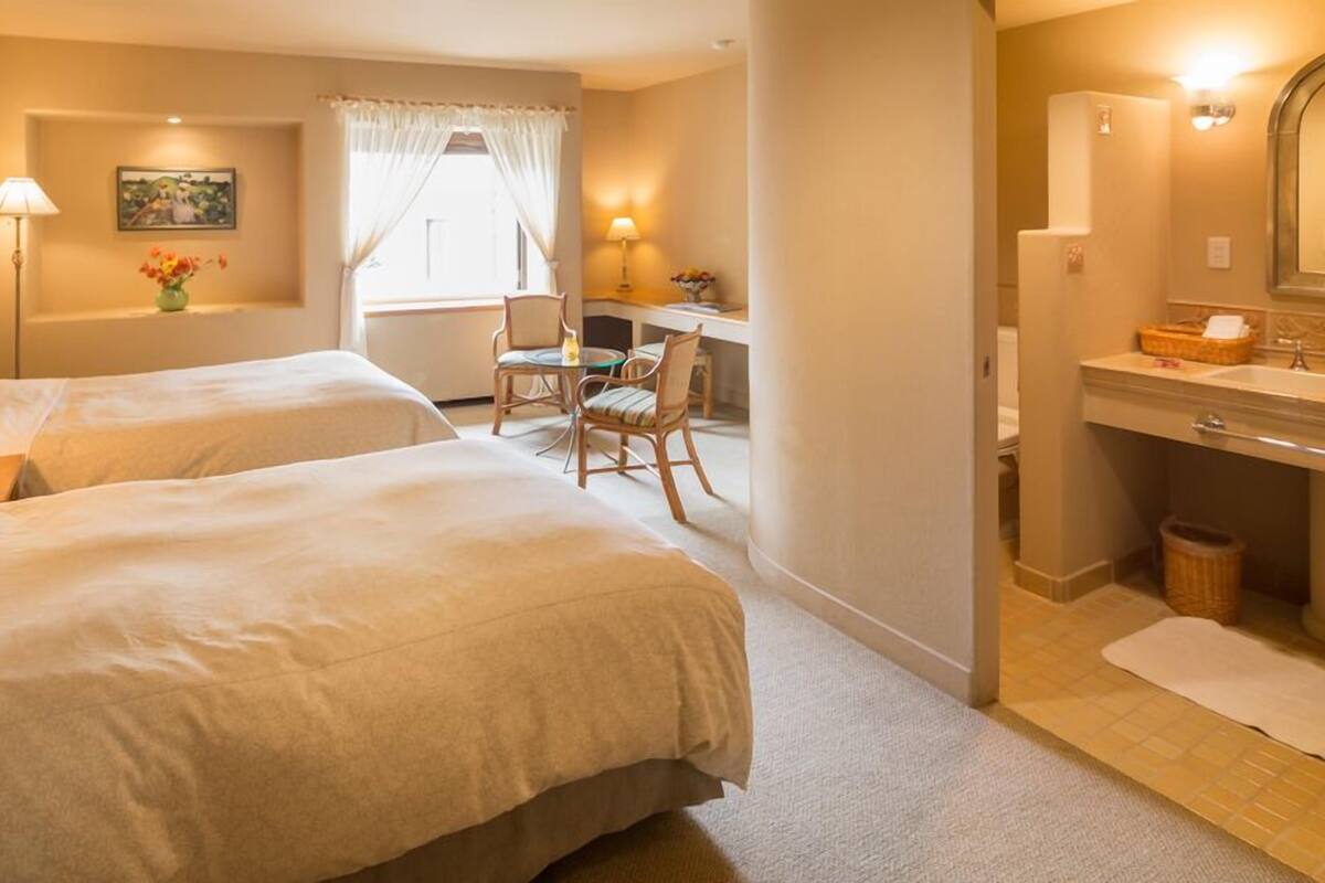 Hotellihuone, jossa on kaksi sänkyä, työpöytä tuolilla, kylpyhuone peilillä sekä ikkuna verhoilla.
