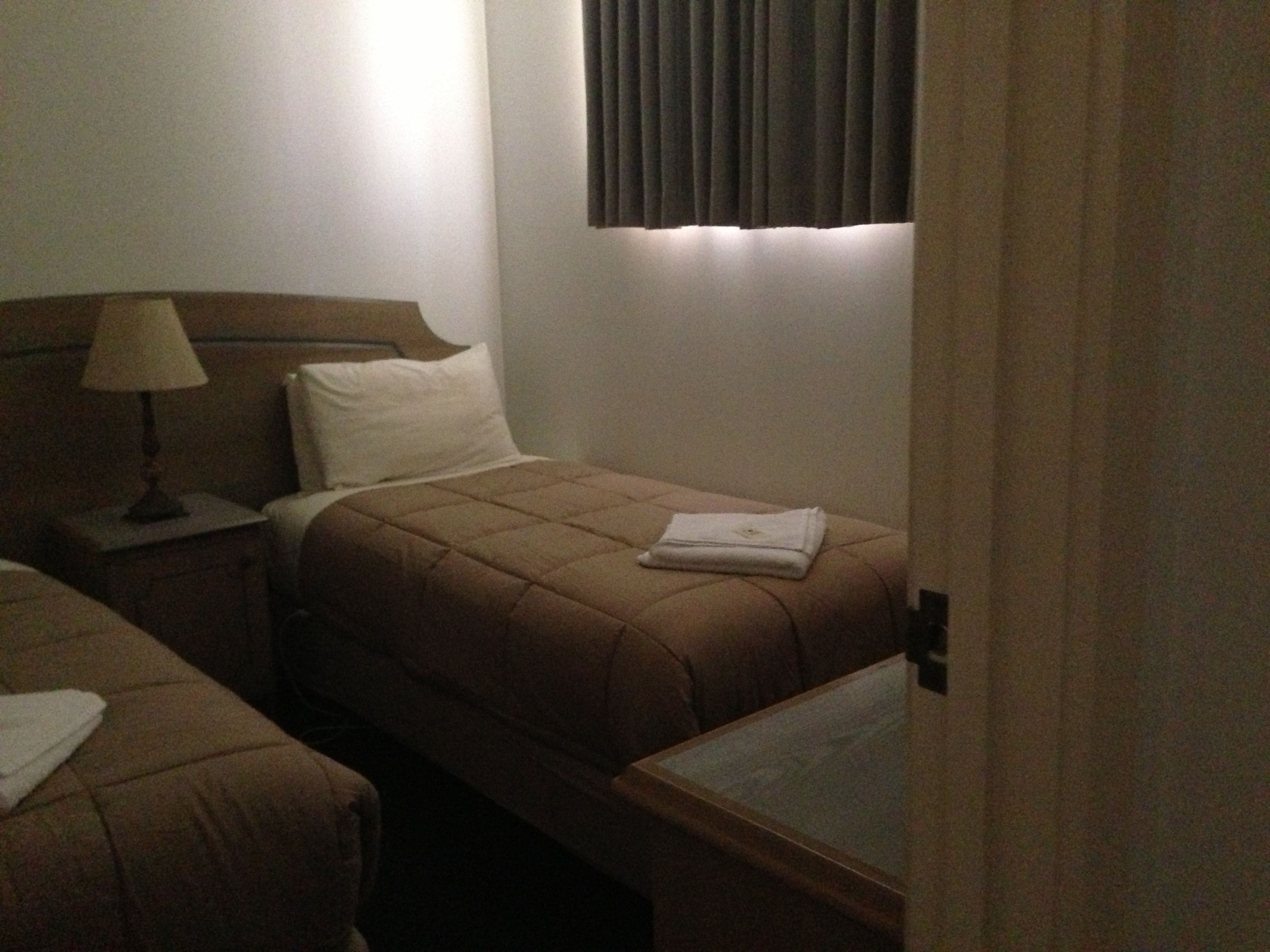 A hotel room with two beds, a bedside table with a lamp, a window with blinds, and a small table with a glass top.
