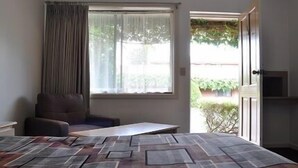 Ein Zimmer mit Bett, Stuhl, kleinem Tisch, einem Fenster mit Vorhängen und einer zur Außenwelt führenden Tür.