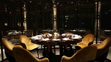 An indoor dining area with a round table set for six, surrounded by four upholstered chairs. The room features a modern design with glass partitions and a view of the cityscape through large windows.