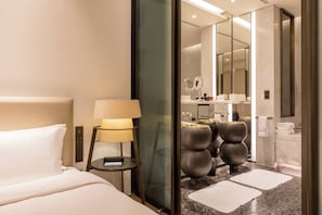 A modern hotel room with a bed, a bedside lamp, a glass partition, and a bathroom visible through the partition.