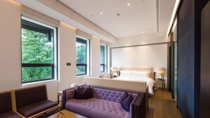 Une chambre d’hôtel moderne, dotée d’un grand lit, d’un canapé, d’une table basse et offrant une vue sur la verdure par les fenêtres.