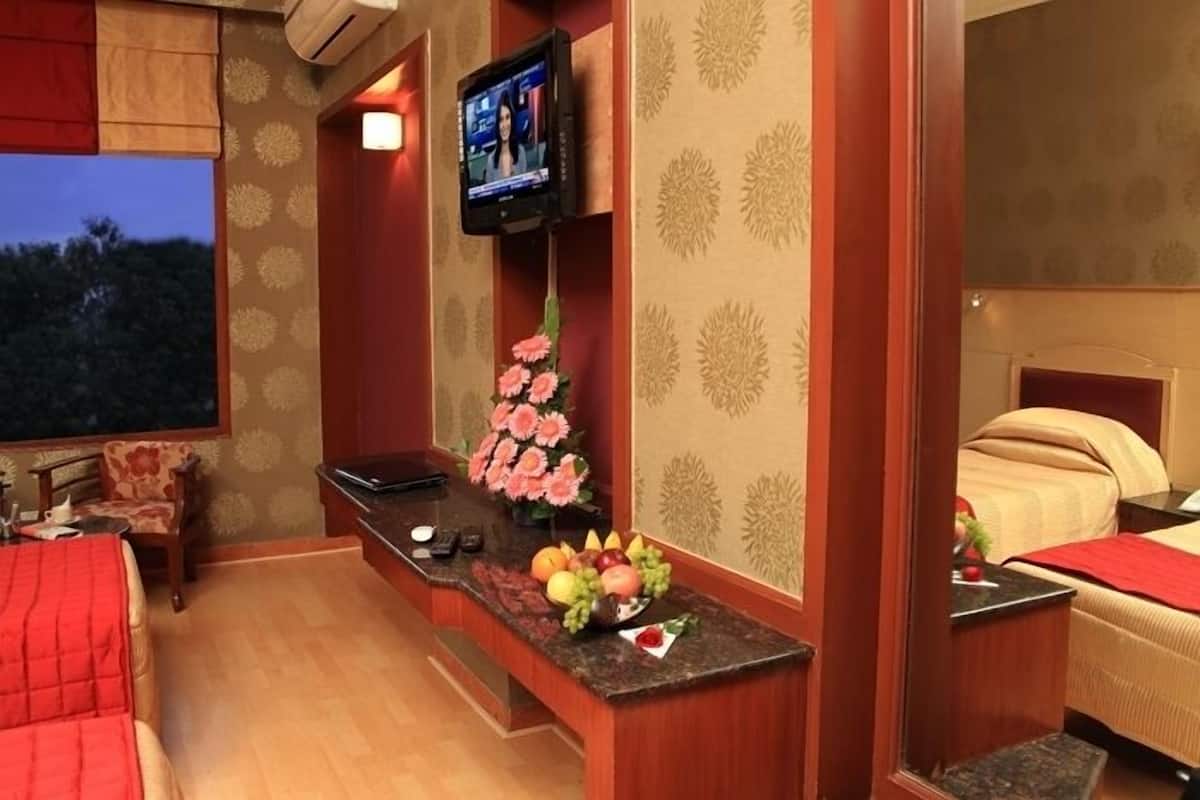 A hotel room with a TV mounted on the wall, a small desk with a vase of flowers, and a bed with a red blanket.